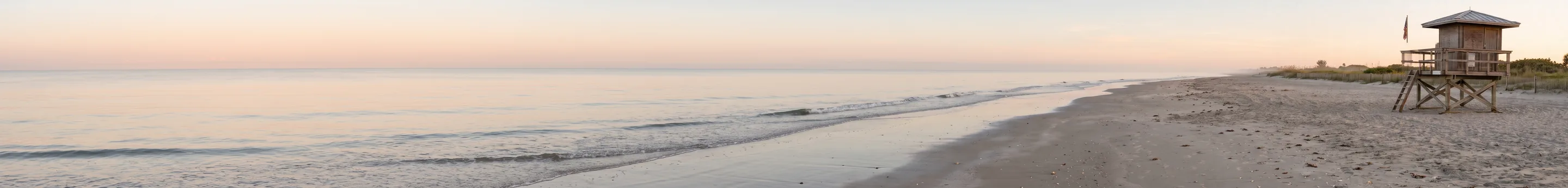 Ein ultrabreiter Streifen eines leeren Strandes bei Sonnenaufgang: blassrosa Himmel und ruhiges Meer über die linken zwei Drittel, nasser Sand, der sich zu einem hölzernen Rettungsschwimmerturm rechts schwingt.