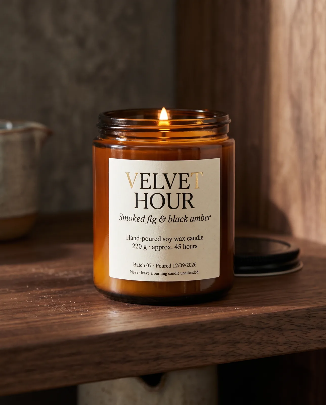 Una candela accesa in un barattolo di vetro ambrato su una mensola di noce scuro. L'etichetta color crema riporta VELVET HOUR, Smoked fig & black amber, Hand-poured soy wax candle, 220 g · approx. 45 hours, Batch 07 · Poured 12/09/2026 e Never leave a burning candle unattended.