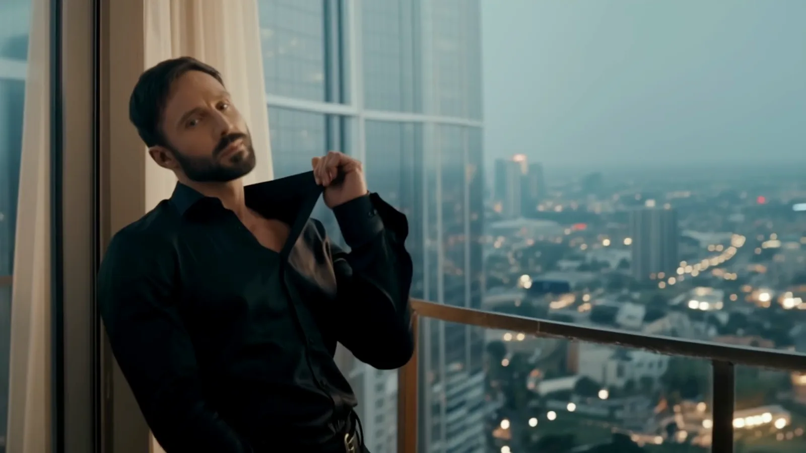 Fotogramma video di un uomo adulto con la barba corta e una camicia di seta nera, appoggiato a un'alta vetrata sopra una città all'ora blu, che si apre il colletto e guarda la camera.