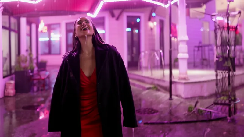 Fotogramma video di una donna adulta con un cappotto scuro sopra un abito rosso che ride con il viso rivolto alla pioggia, sotto un portico illuminato da un neon rosa, di notte.