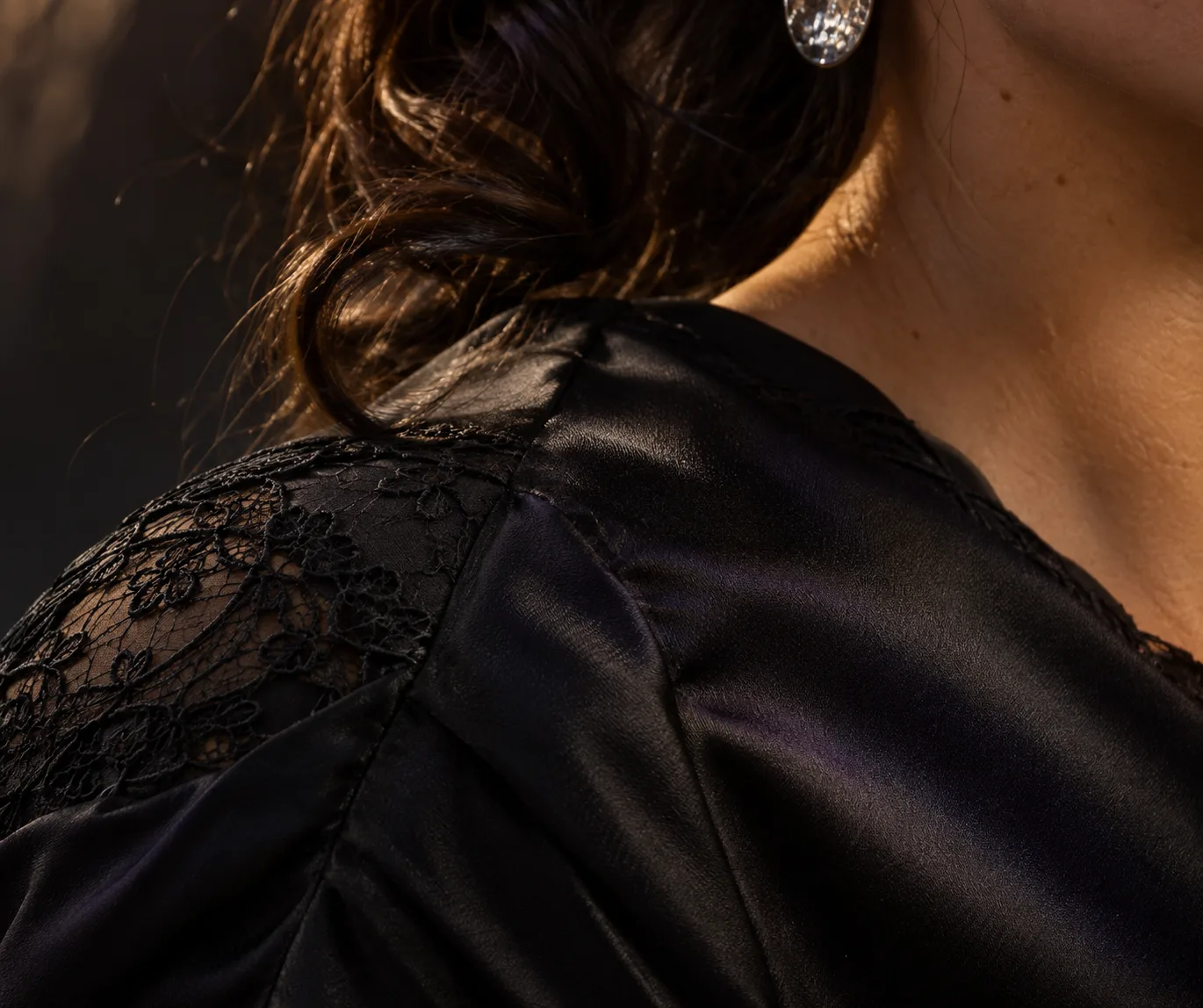 Un dettaglio di moda mostra una cucitura sulla spalla in satin nero, decorazione in pizzo, ricci raggruppati e orecchino argento sotto luce laterale radente.