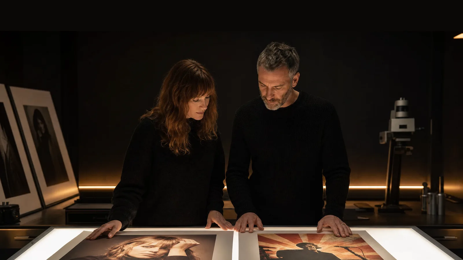 A woman with auburn hair and a bearded man in dark knitwear comparing two large photo prints on a backlit table in a dim studio