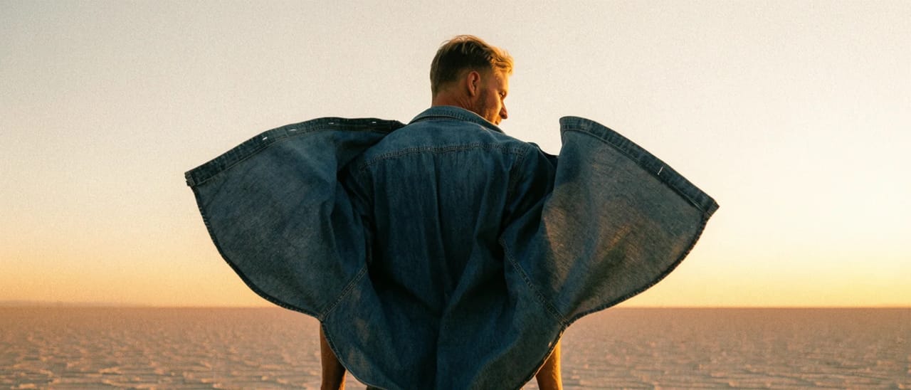 A man seen from behind on a salt flat at sunrise, his denim shirt blown out wide on both sides like wings.