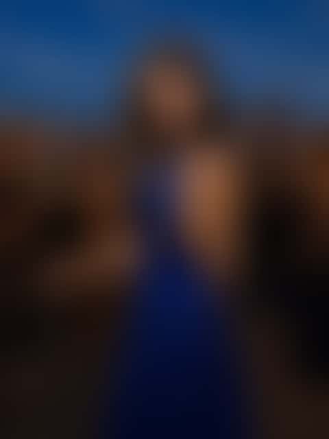 A woman in a royal-blue sequined halter gown stands with one hand on the railing at a blue-hour rooftop party, string lights overhead and guests with glasses in front of a glittering skyline