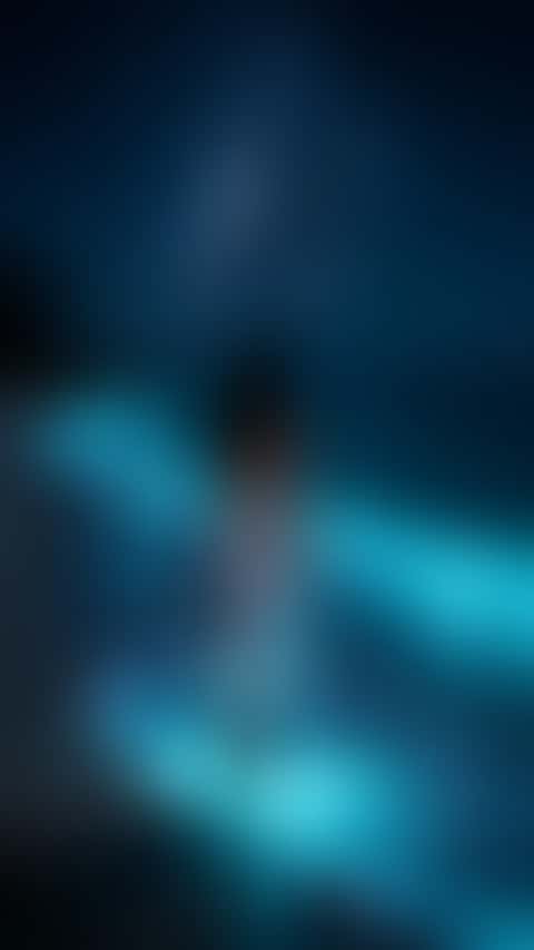 On a night beach under the Milky Way, a woman in white silk wades into waves glowing electric blue and looks back over her shoulder with a smile