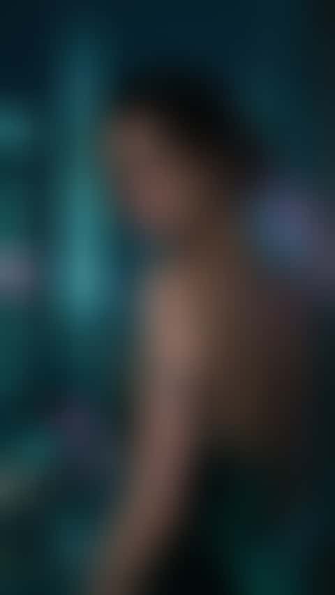 A woman in her thirties in a deep emerald velvet dress leans at a rain-streaked night window, pink and teal neon glowing on the glass and her collarbone as she glances back with a half smile.