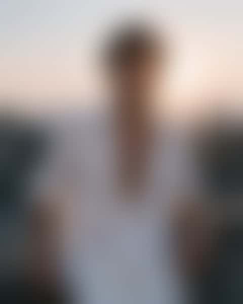 A man in his twenties stands on a dusk concrete rooftop in an open white linen shirt with rolled sleeves, one arm along the railing, backlight tracing his jaw and chest against a hazy skyline.