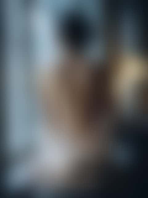 A woman seen from behind at a tall window, champagne satin draped low around her back, in cold morning light.