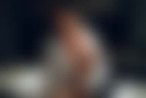 A woman with wet hair in an oversized white shirt slides off a dark kitchen counter and walks toward the camera, a wine glass and a steaming pan behind her.