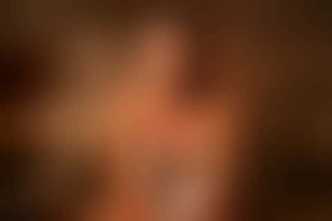 A woman with a towel sits in a wooden sauna, tips a ladle of water onto the stones, vanishes in a burst of steam and wipes her collarbone looking at the lens