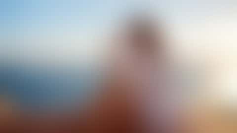 Una donna seduta sul parapetto di un tetto mediterraneo all’ora d’oro, in camicia di lino bianca aperta, si alza gli occhiali da sole sui capelli e ride, con il mare alle spalle