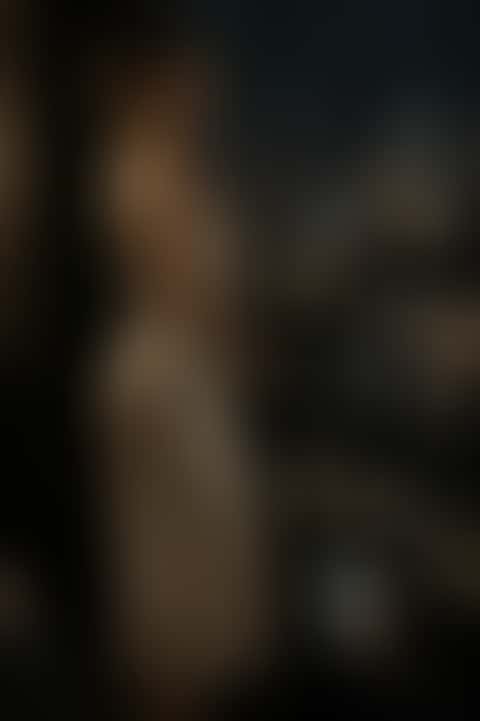 A woman in champagne satin by a rain-streaked window at night turns to face the camera