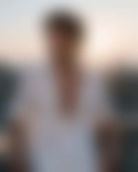 A young man on a concrete rooftop at dusk turns from the railing as the wind lifts his open white linen shirt, then runs a hand through his hair.