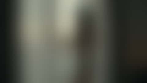A woman in a short ivory silk chemise stands at a window with sheer curtains, turns slowly toward the lens and lifts her eyes to the camera