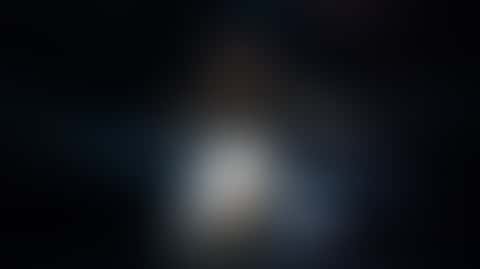 A dancer in layered grey-white chiffon spins on a dark stage as a follow spot from behind turns the skirt translucent and glowing