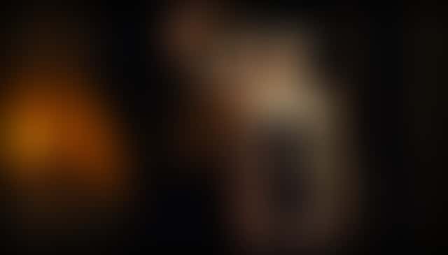 A woman in a black lace bodysuit seen from behind, her front reflected in a tall antique mirror in a lamp-lit bedroom