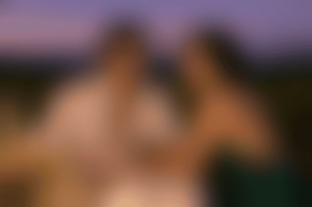 A man in an open white shirt and a woman in a backless emerald silk dress lean toward each other at a candlelit table on a vineyard terrace at dusk