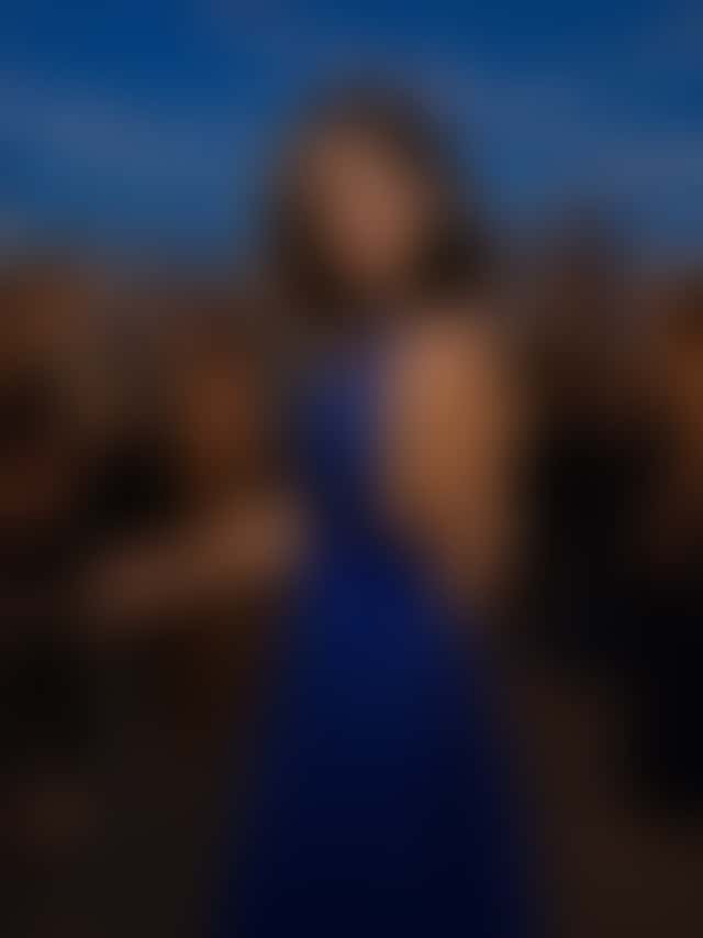 A woman in a royal-blue sequined halter gown stands with one hand on the railing at a blue-hour rooftop party, string lights overhead and guests with glasses in front of a glittering skyline