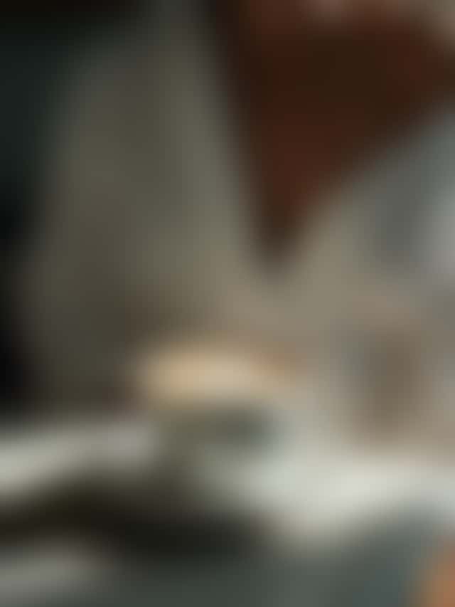 A latte on a sunlit white kitchen counter is lifted out of frame as the camera tilts up to a woman who smiles softly at the viewer