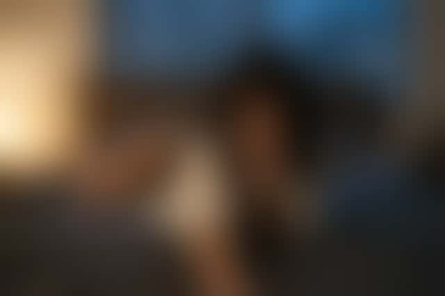 Una donna con una camicia bianca aperta si sporge su un letto scuro verso la camera, un dito sul labbro, poi accenna un lento mezzo sorriso; dietro, una lampada e una finestra blu.