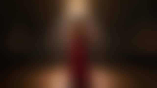 In una sala da ballo vuota sotto un lampadario di cristallo, un uomo in gilet nero inclina all’indietro sul suo braccio una donna in abito di raso rosso, e i due si chinano l’uno verso l’altra, fronte contro fronte.