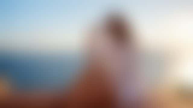 Una donna seduta sul parapetto di un tetto mediterraneo all’ora d’oro, in camicia di lino bianca aperta, si alza gli occhiali da sole sui capelli e ride, con il mare alle spalle