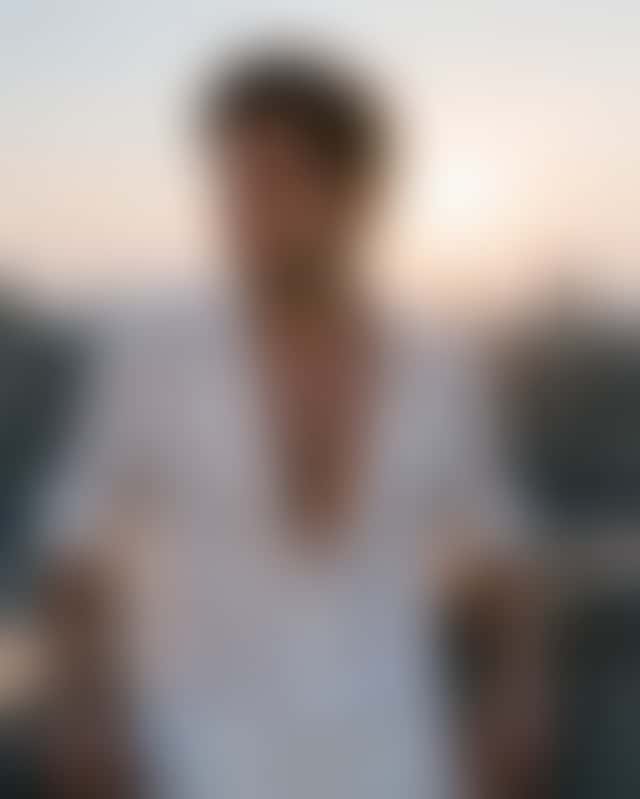 A young man on a concrete rooftop at dusk turns from the railing as the wind lifts his open white linen shirt, then runs a hand through his hair.