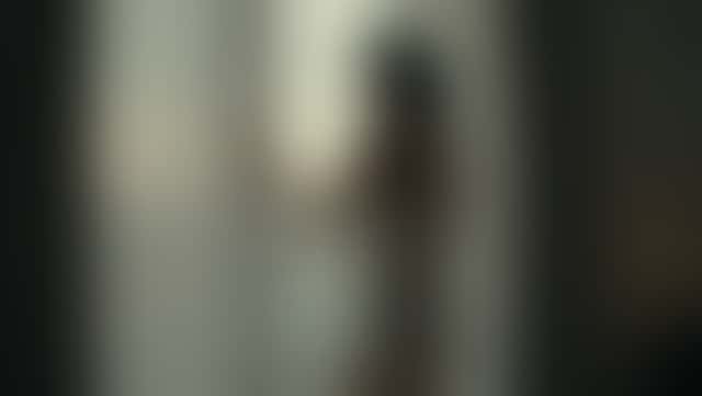 A woman in a short ivory silk chemise stands at a window with sheer curtains, turns slowly toward the lens and lifts her eyes to the camera