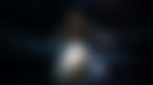 A dancer in layered grey-white chiffon spins on a dark stage as a follow spot from behind turns the skirt translucent and glowing