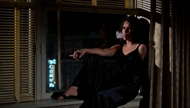 Night interior: a woman on an apartment windowsill exhaling smoke against rain-streaked glass while a red neon sign pulses across her face