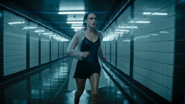 The camera retreats down a wet, blue-lit pedestrian tunnel as a woman in a translucent raincoat over a black dress runs toward it and pushes open a steel door