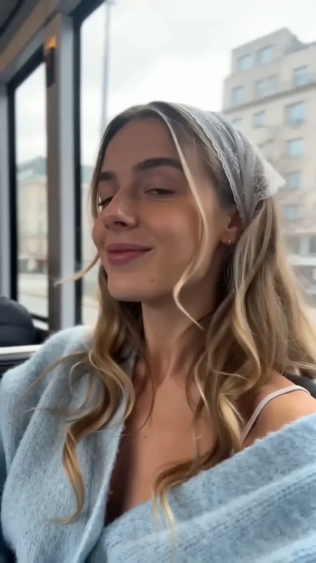 A vertical phone-camera clip of a woman by a city bus window in a pale blue cardigan, winking and flashing a peace sign