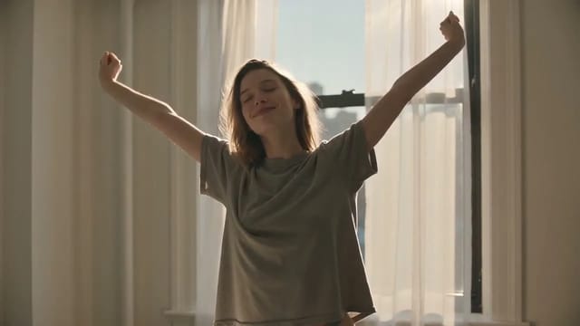 A woman in an oversized grey t-shirt stretching beside a sunlit window in a studio apartment, then flopping down laughing