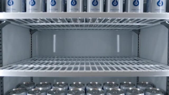 A locked shot of a drinks fridge shelf where a gloved hand fills the empty row with silver cans one by one, labels facing forward