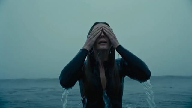 A woman in a black wetsuit stands in grey, foggy surf at dawn, wrings seawater out of her hair, tips her head back and smiles into the lens as a wave breaks behind her