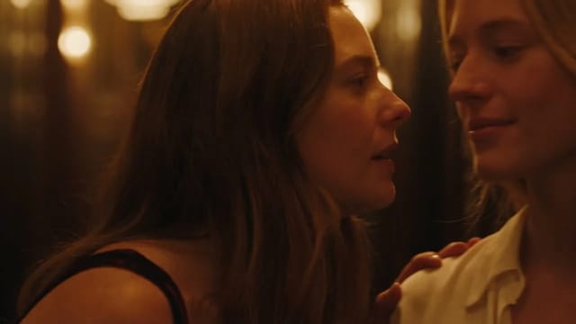 Two women standing close together in the amber light of a jazz bar hallway, one whispering at the other's ear while she laughs