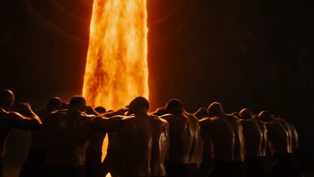 Molten iron pours down in a vast dark forge, lighting a long row of smiths in orange as they shield their faces from the heat