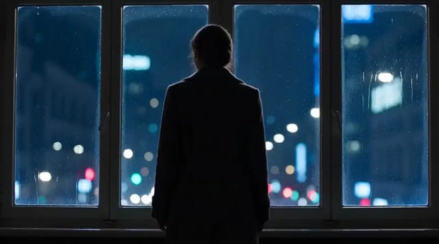 A woman in a long dark coat stands at a wall of rain-streaked windows at night, city lights blurred beyond, and turns her head back over her shoulder.