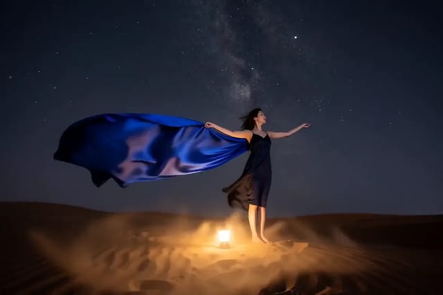 A woman in a dark slip dress throws a huge sheet of indigo silk overhead on a desert dune at night, a lantern at her feet and the Milky Way above.