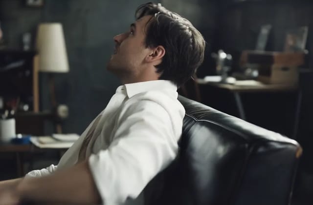 A man in an open white shirt leans back into a leather sofa in a dim office and turns his head toward the camera.