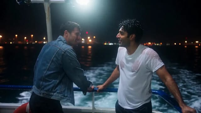 Two men at the stern rail of a night ferry, one in an open denim jacket and one in a soaked white T-shirt, pressing their foreheads together laughing before they kiss, the wake churning white below.