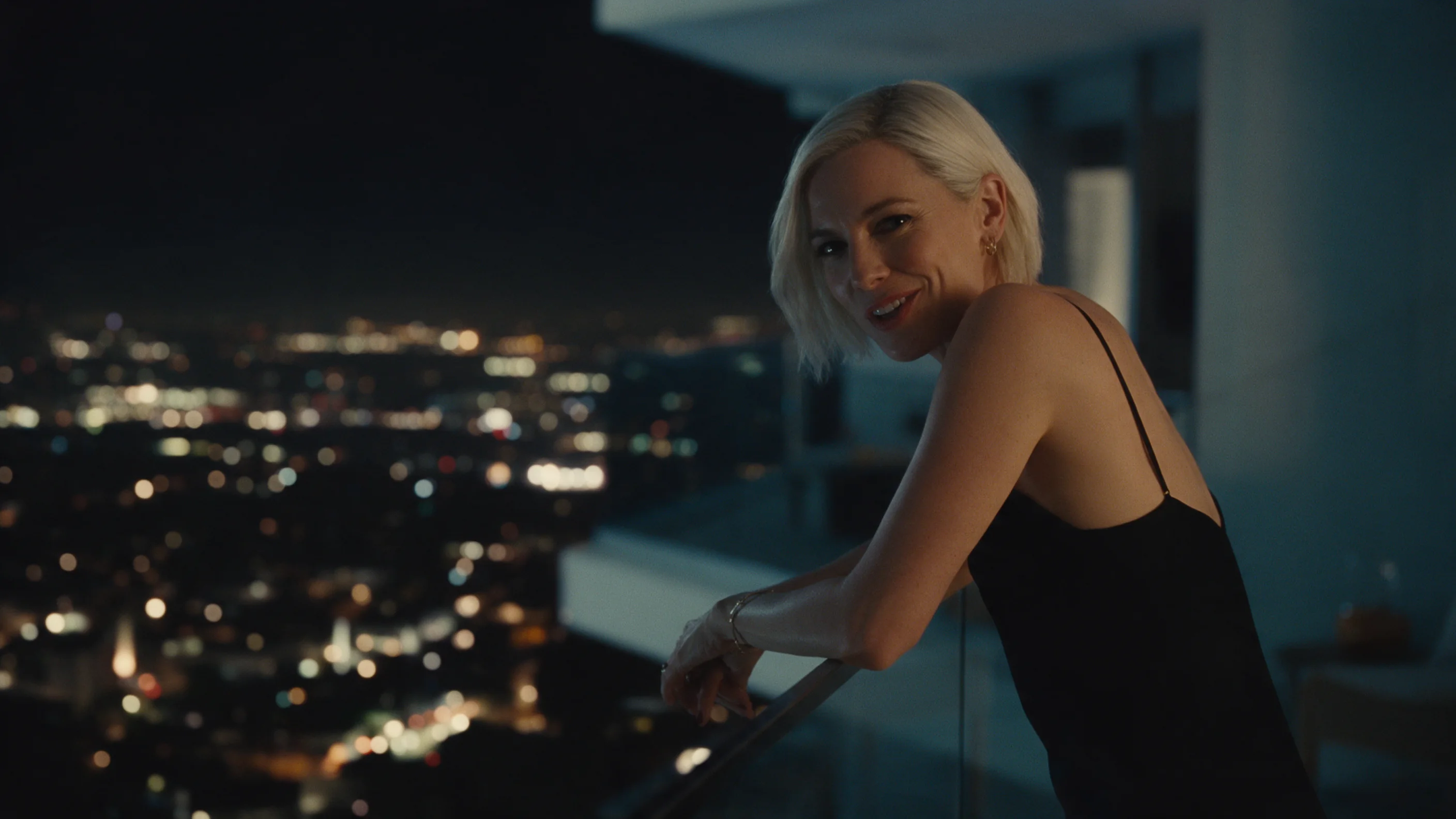 Image fixe cinématographique d’une femme au carré court blond platine, en robe noire en satin à fines bretelles, accoudée à la rambarde en verre d’un balcon, la nuit : elle se tourne vers la caméra en pleine phrase, avec un demi-sourire malicieux, les lumières de la ville floues en contrebas.
